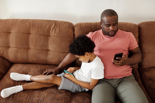 man sitting on couch looking down at cellphone with young child sitting sideways on the couch while leaning against him