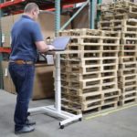 worker standing at laptop on stand with band turned away from camera, standing in front of storage shelves and wooden pallets