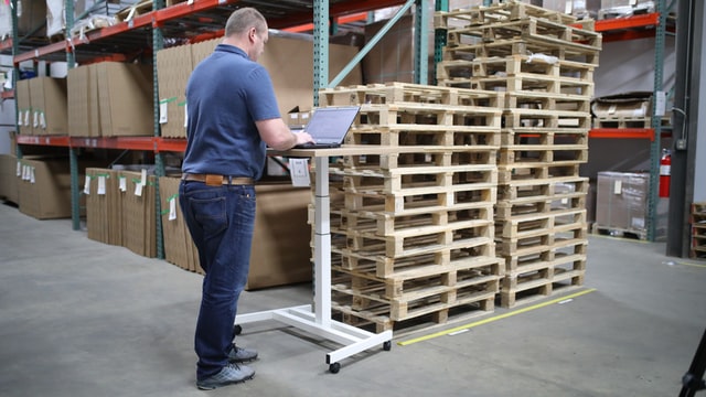 worker standing at laptop on stand with band turned away from camera, standing in front of storage shelves and wooden pallets